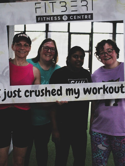 Four women holding a sign at a Fitberi Fitness Center, with text 'I just crushed my workout'.